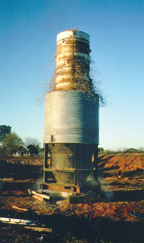A large cylindrical soil testing apparatus ejects gravel and debris during a geotechnical experiment at an outdoor construction site, with trees and dirt piles in the background.